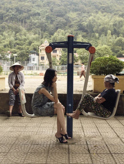 Three women seated on outdoor fitness equipment in a park setting with a lush green mountain backdrop and nearby buildings.