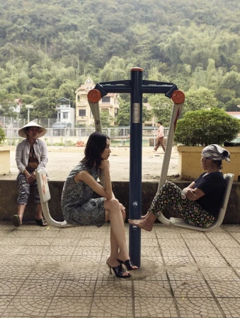 Three women seated on outdoor fitness equipment in a park setting with a lush green mountain backdrop and nearby buildings.