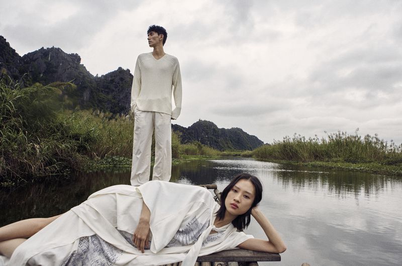 Two fashion models in off-white clothing pose on a bamboo raft along a narrow river, set against a backdrop of dramatic mountains and a cloudy sky.