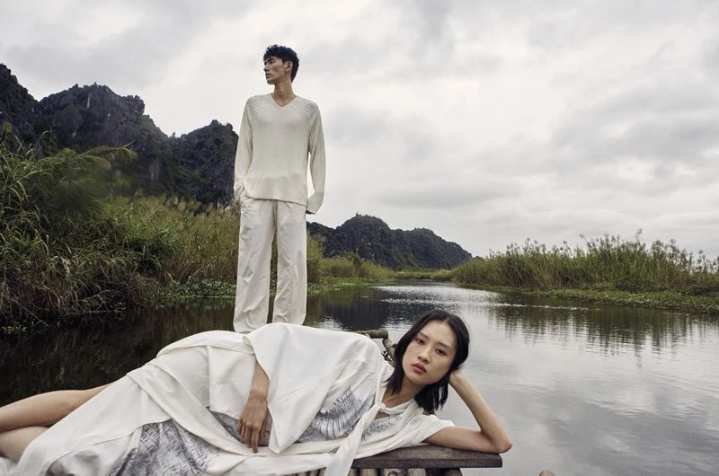 Two fashion models in off-white clothing pose on a bamboo raft along a narrow river, set against a backdrop of dramatic mountains and a cloudy sky.
