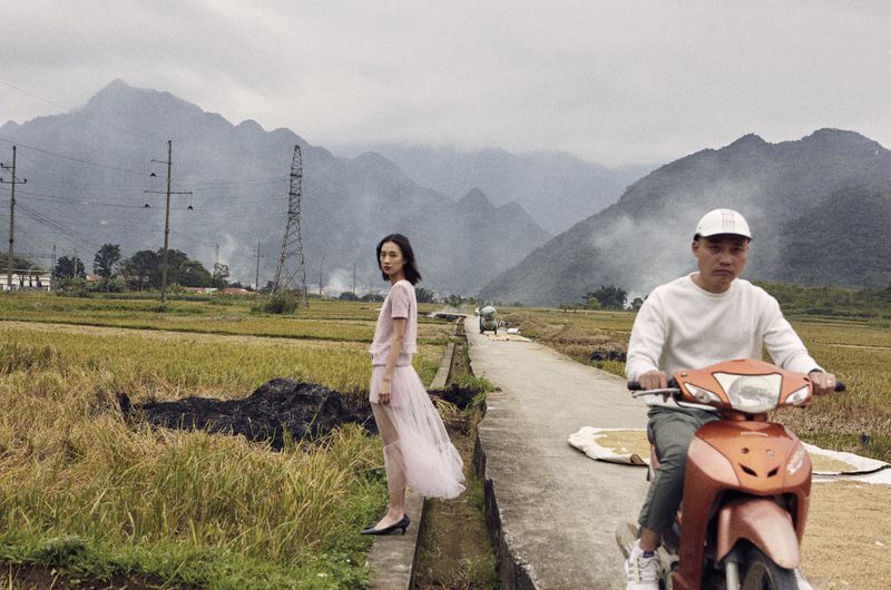 A woman in a pink outfit stands near rice paddies, while a man drives a moped on a rural road under an overcast sky in a fashion editorial.