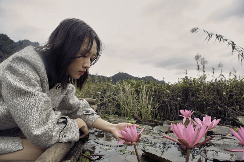 Young woman in gray coat touches a pink water lily in a pond surrounded by green vegetation and distant mountains under an overcast sky.