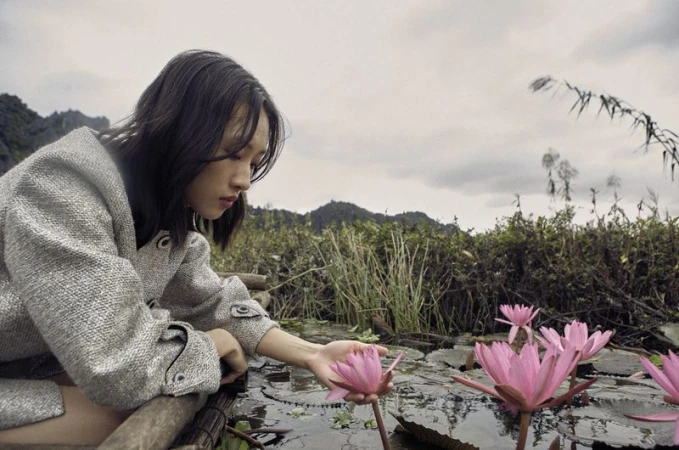 Young woman in gray coat touches a pink water lily in a pond surrounded by green vegetation and distant mountains under an overcast sky.