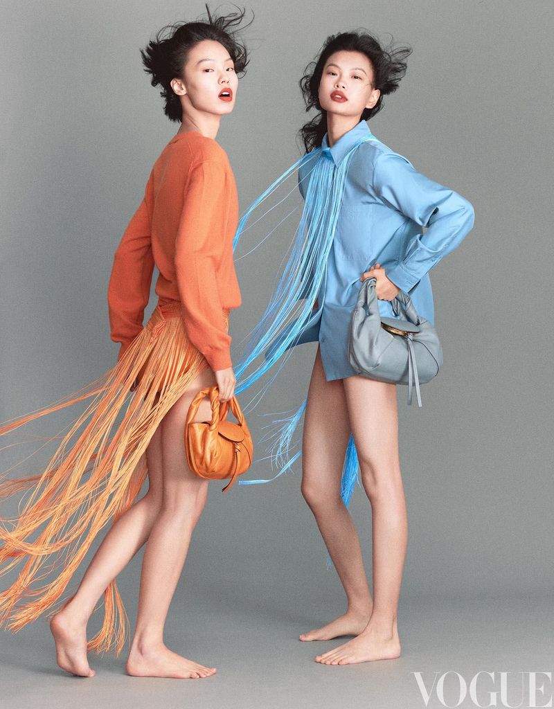 Two women in fashionable outfits pose in a studio, one in orange with a fringed skirt, the other in blue with a purse, showcasing vogue style.
