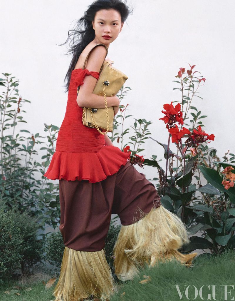 Editorial fashion shot of an Asian model in a red dress and brown pants with straw fringe, holding a purse, outdoors on a grassy hill.
