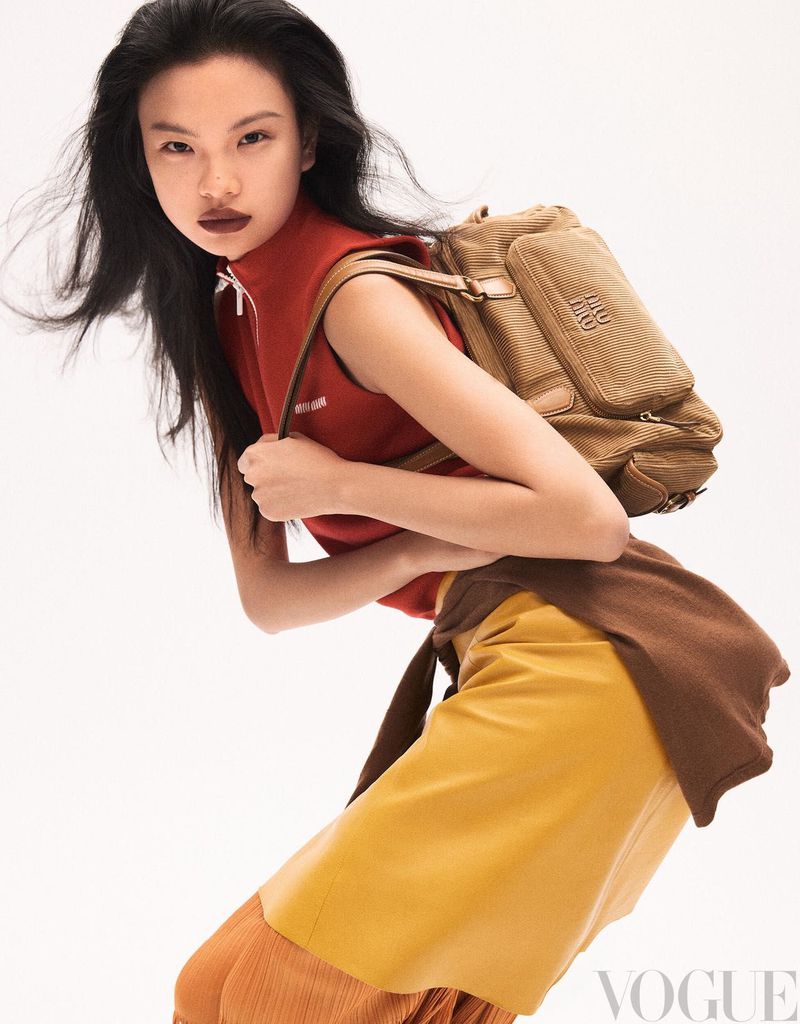 Fashion editorial shot of an Asian woman with long dark hair, wearing a red top, yellow skirt, and backpack against a white backdrop.