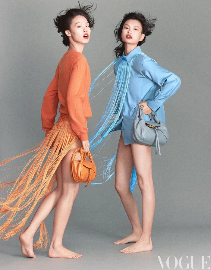 Two female models posing in colourful fringed tops and shirts carrying handbags against a grey studio background for Vogue China.