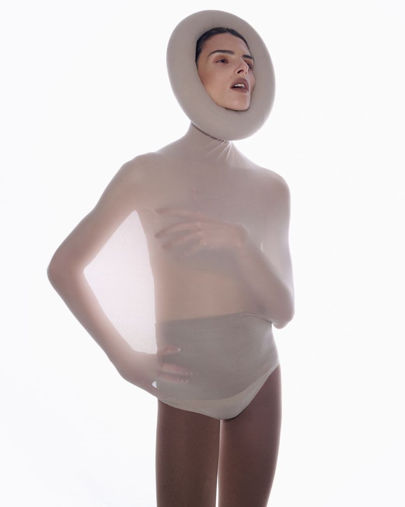 Woman in nude bodysuit and round headpiece poses in bright studio. Fashion, artistic portrait, nude, and headpiece elements.