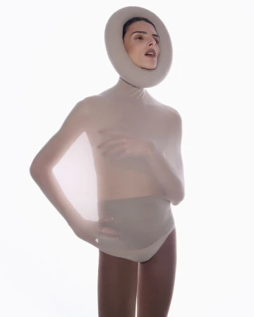 Woman in nude bodysuit and round headpiece poses in bright studio. Fashion, artistic portrait, nude, and headpiece elements.