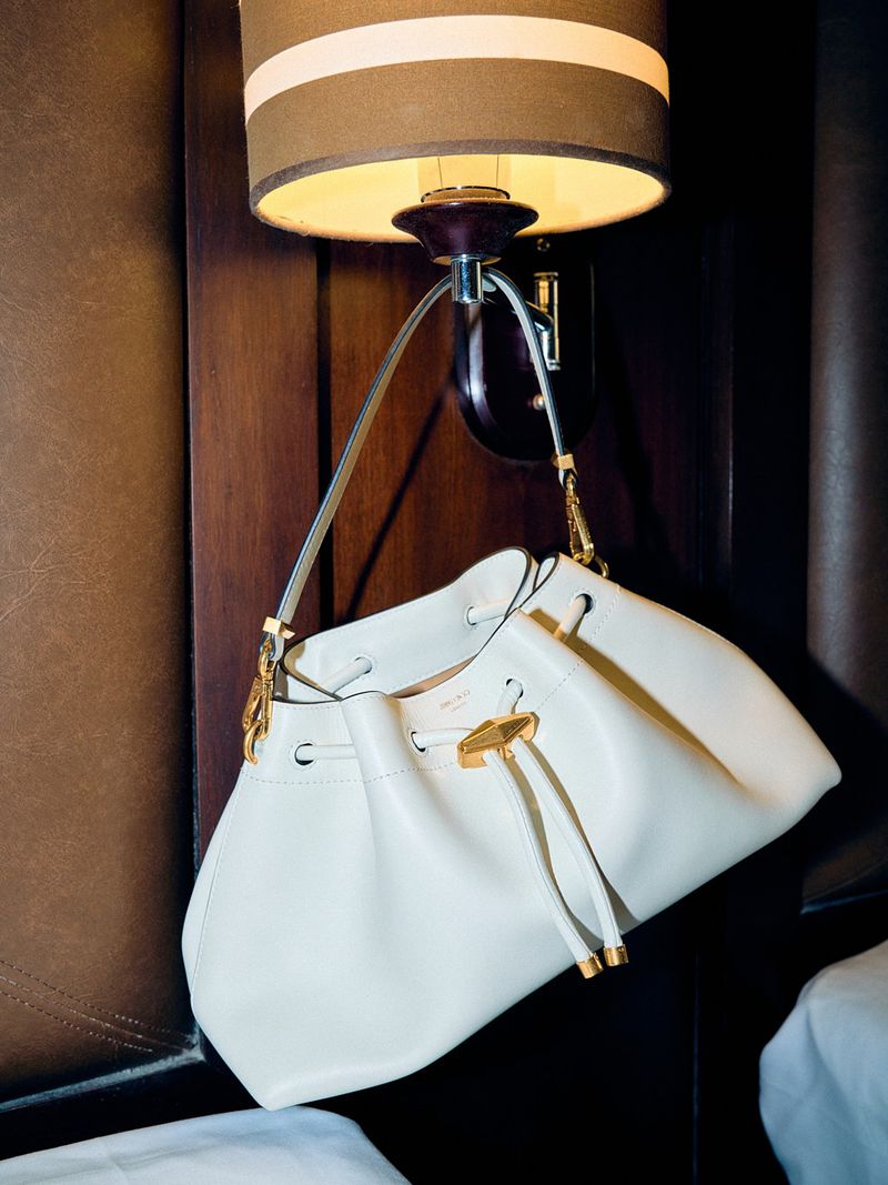 Elegant white leather bucket bag with gold hardware hanging from a wall lamp against a brown leather headboard backdrop.