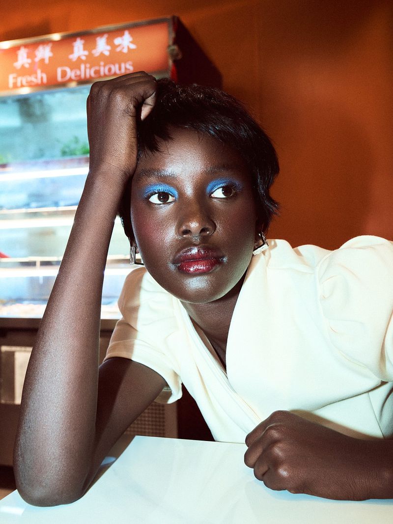 Close-up of a young woman with bold blue eyeshadow, wearing a cream-colored top, sitting at a table in an indoor setting.
