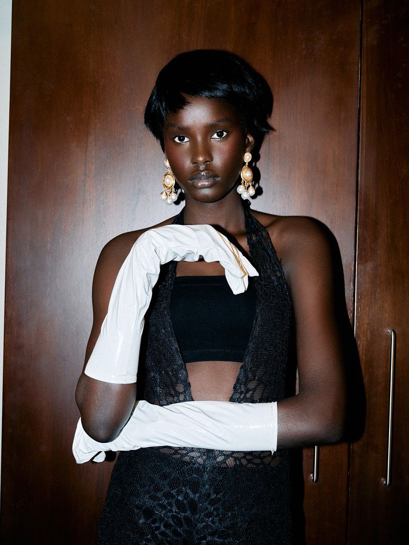 Elegant woman with dark skin poses in black lace top and white gloves, accessorized with pearl earrings, against wood panel background.