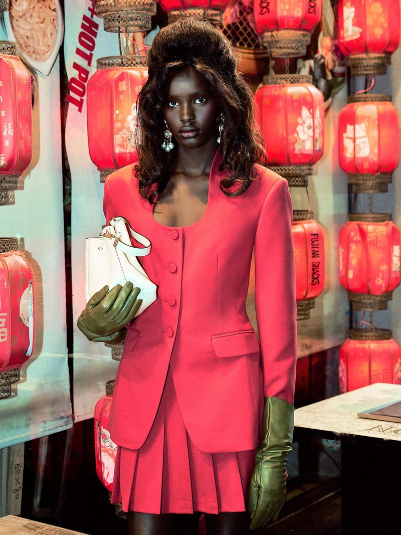 Fashionable woman in a pink skirt suit and gloves, holding a white purse, standing indoors with decorative red Chinese lanterns.