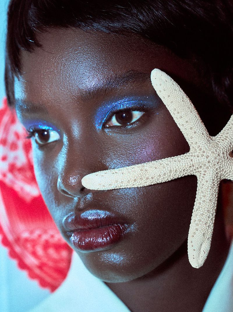 Close-up fashion portrait of a woman with dark skin, glossy lips and vivid blue eye makeup with a starfish near her face.