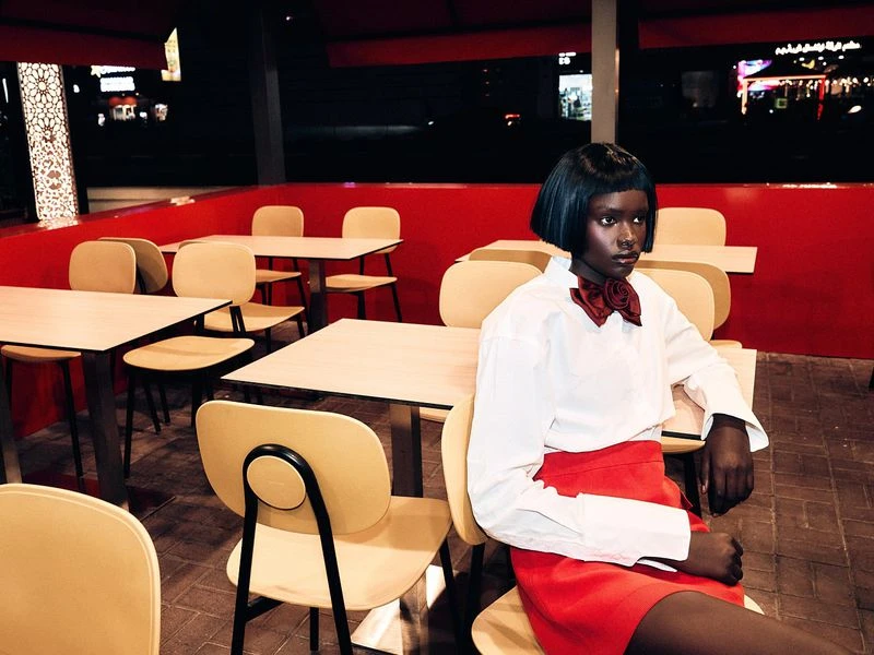 Elegant dark-skinned woman in a red skirt and white blouse poses in a restaurant setting with tan chairs and red accents at night.