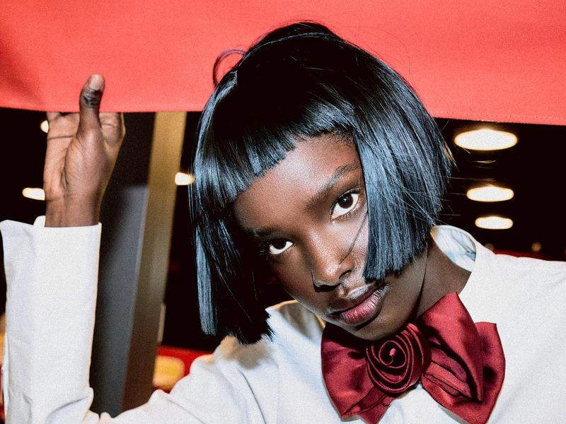 Portrait of an African American woman with short bob hair and a red bow tie, posing in front of a dark background with bright lights.