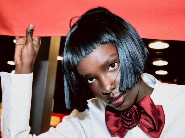 Portrait of an African American woman with short bob hair and a red bow tie, posing in front of a dark background with bright lights.