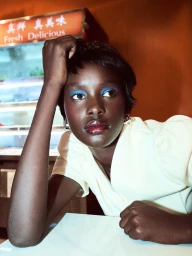 Model Abuk Mayen with vibrant blue eyeshadow and glossy lips resting her head on her hand at a white table.