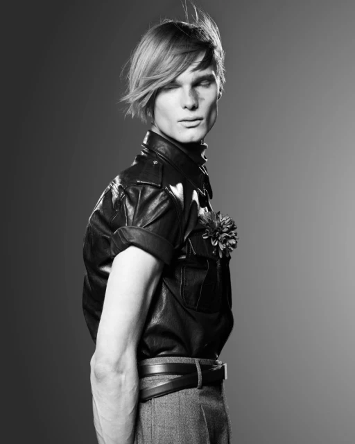 Monochrome studio portrait of a slender adult male model with styled blonde hair wearing a leather shirt and grey pants against a grey background.