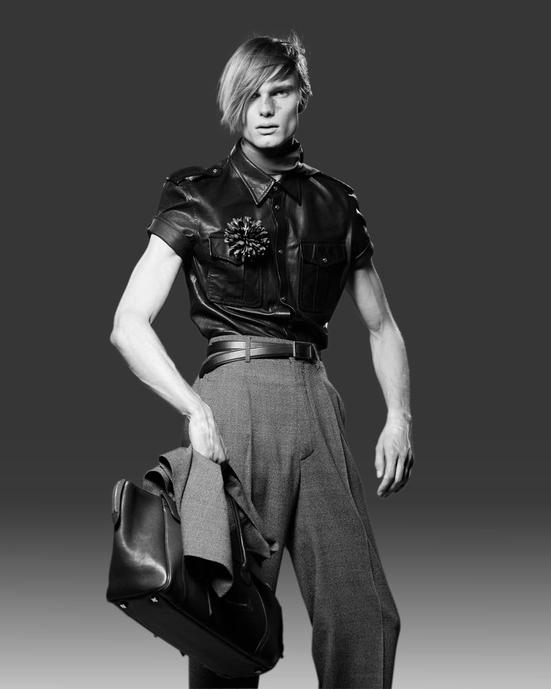 Black and white studio shot of a male fashion model wearing a leather shirt and grey trousers, carrying a suit jacket and bag.
