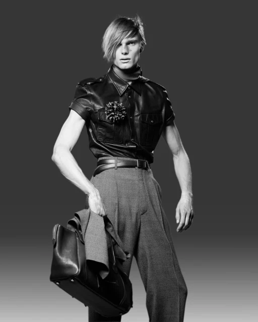 Black and white studio shot of a male fashion model wearing a leather shirt and grey trousers, carrying a suit jacket and bag.