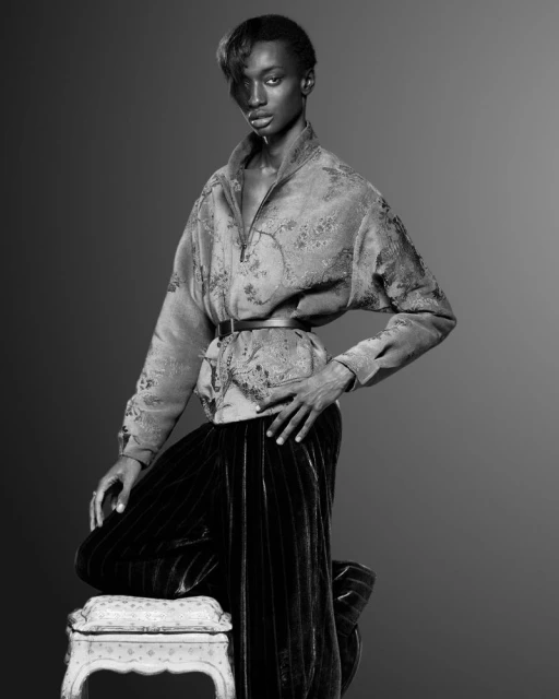 Monochrome portrait of a black, adult woman in a patterned jacket, velvet pants, and heels, kneeling on a stool in a photo studio.