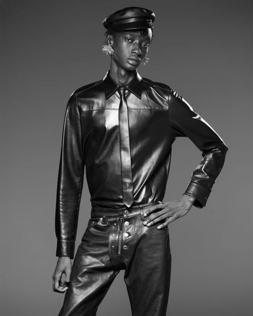 Monochrome fashion portrait of a slender person in black leather shirt, pants, tie, and cap against a dark gray seamless background.