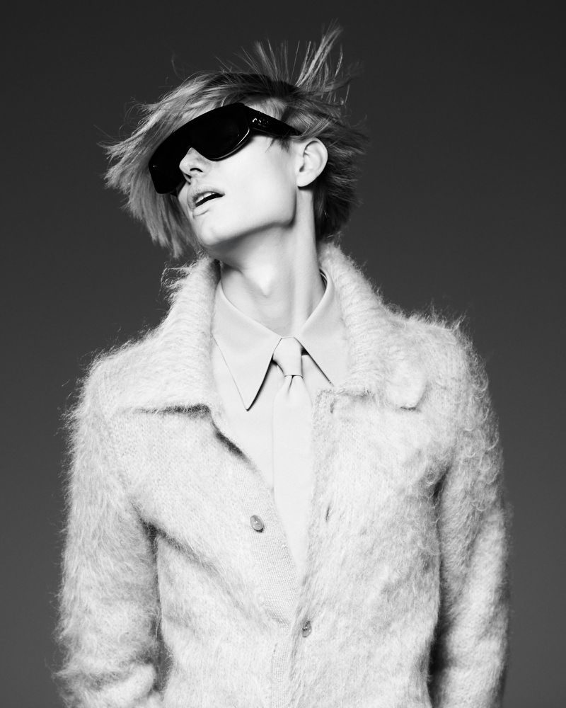 Black and white portrait of a person with spiky blond hair wearing a fuzzy coat and large sunglasses against a solid neutral backdrop.