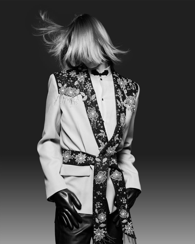 Fashion shot of a person wearing a cream colored blazer with floral embellishments and black gloves against a dark gray background.