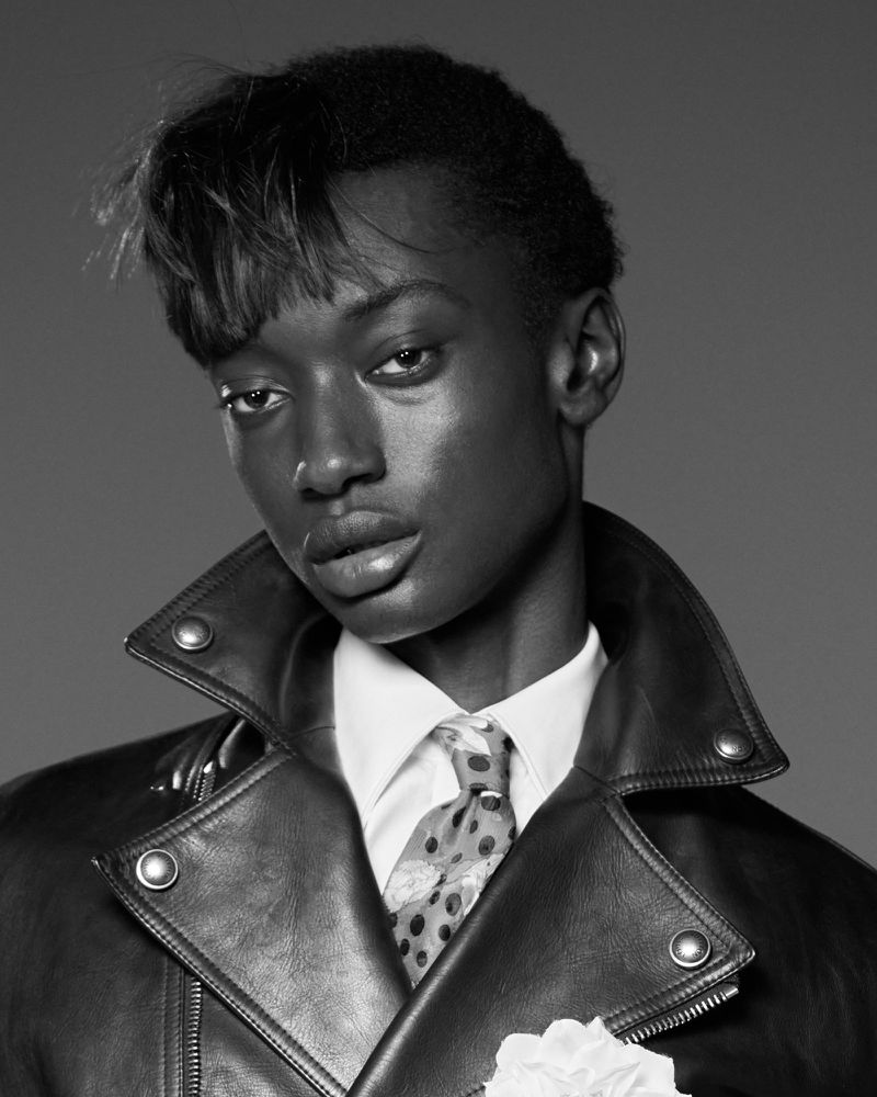 Monochrome portrait of a Black male model in a leather jacket and white collared shirt with a patterned tie and boutonniere.