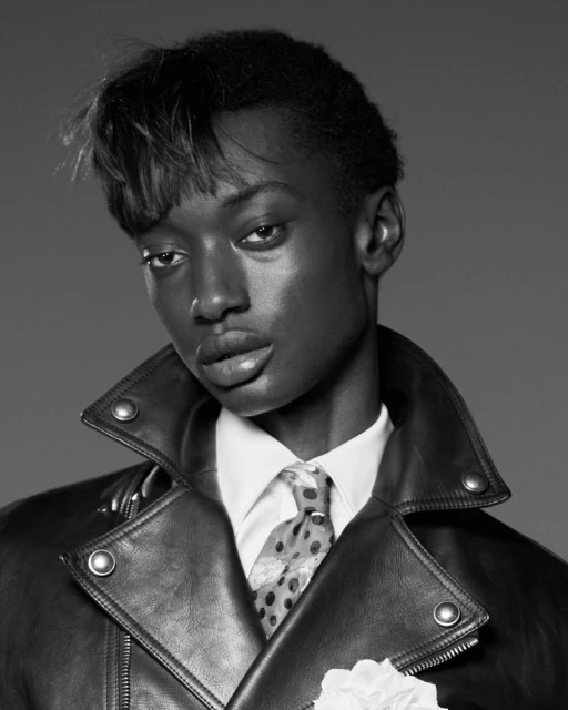 Monochrome portrait of a Black male model in a leather jacket and white collared shirt with a patterned tie and boutonniere.