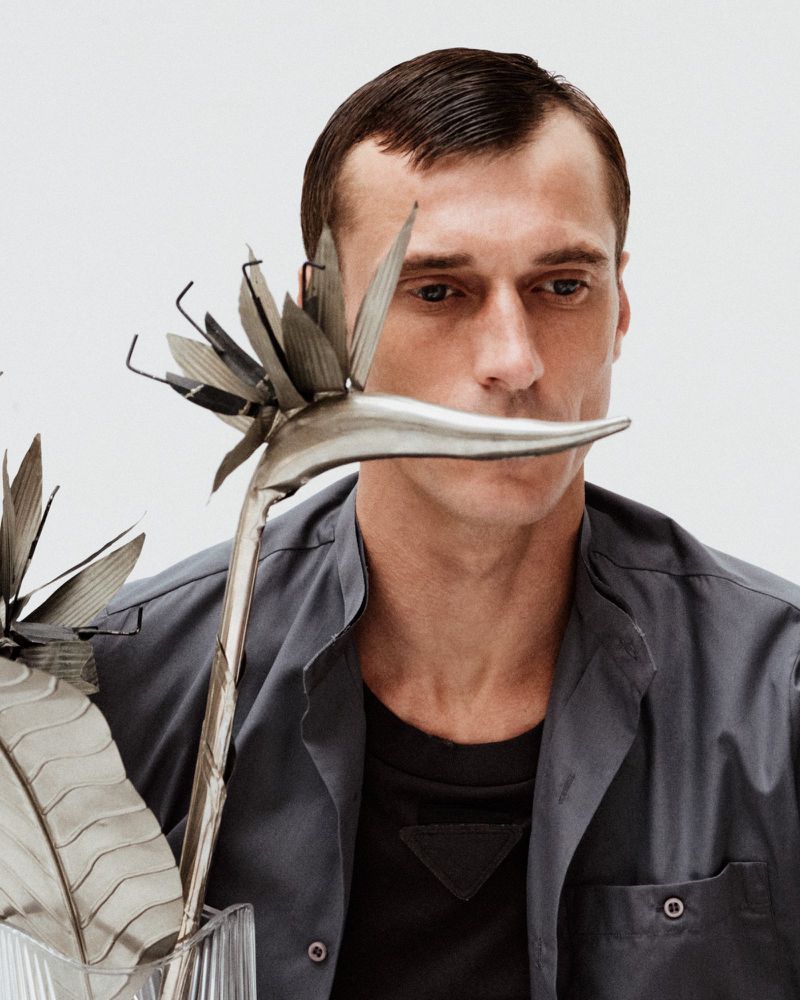Man with short dark hair poses next to two metal bird of paradise sculptures in a studio, wearing an open gray collared shirt.