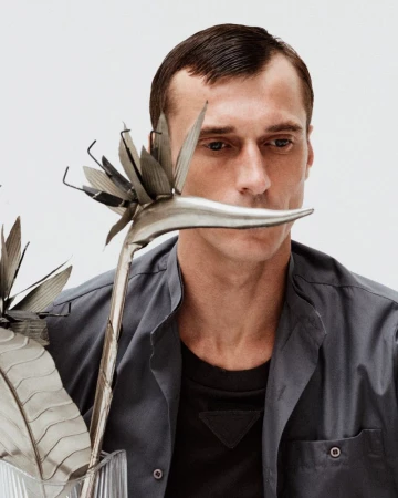 Man with short dark hair poses next to two metal bird of paradise sculptures in a studio, wearing an open gray collared shirt.