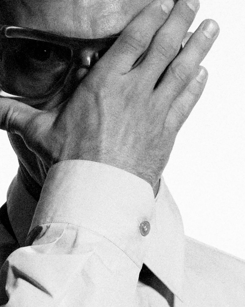 Monochrome portrait of a light-skinned adult man covering face with hand, wearing sunglasses and a button-down shirt against a plain background.