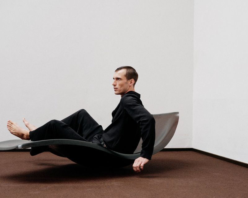 Adult male in a black outfit reclines on a modern grey rocking chair in an indoor setting with white walls and a brown carpet.