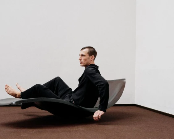 Adult male in a black outfit reclines on a modern grey rocking chair in an indoor setting with white walls and a brown carpet.