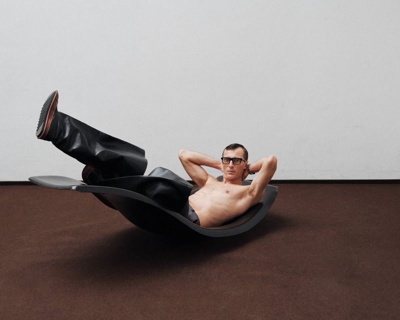 Shirtless man in glasses reclines on a black chair with his hands behind his head in a minimalist interior with a brown floor.