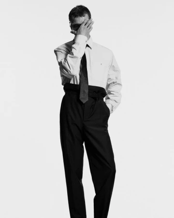 Adult male in a light shirt, dark tie, and dark trousers with one hand covering part of his face in a full studio shot.