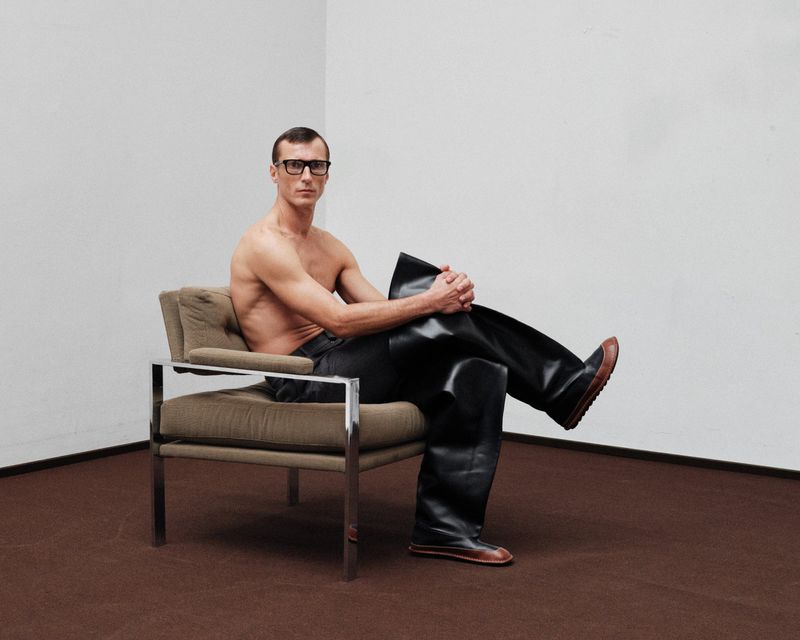 An adult man with bare torso and glasses sits in a chair, wearing shiny black waders, indoors against a plain white wall and brown floor.