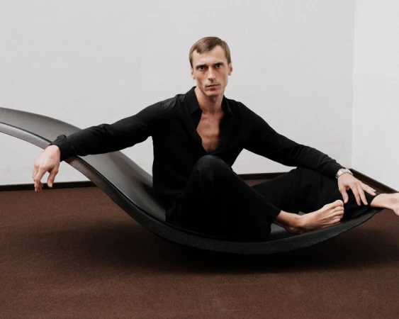 An adult male in a black long-sleeve shirt and pants sits on a modern black lounge chair in a minimalist room with brown carpet.