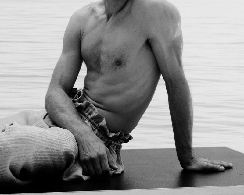 Monochrome portrait of shirtless man with patterned pants seated on dark surface against light rippled water background. 