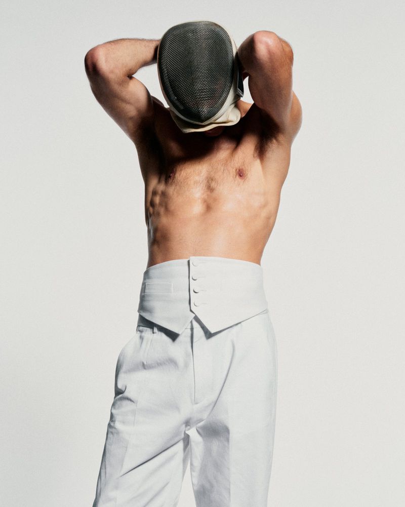 Man in white pants and fencing mask against a white background. Upper body visible, muscular build, and arms behind his head.