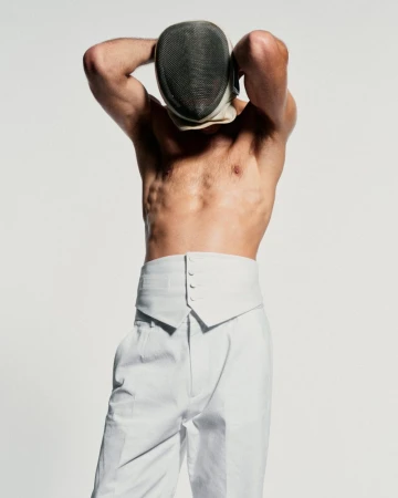 Man in white pants and fencing mask against a white background. Upper body visible, muscular build, and arms behind his head.