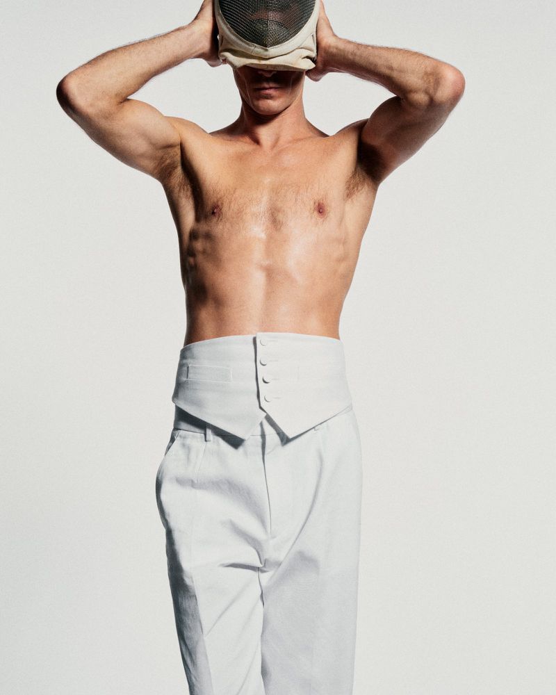 Athletic adult male in high-waisted white trousers and a fencing mask posing against a white backdrop in a studio setting.
