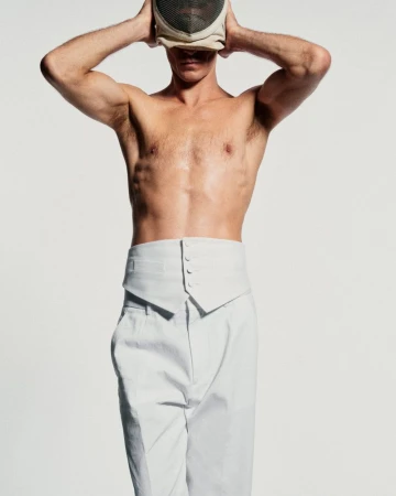 Athletic adult male in high-waisted white trousers and a fencing mask posing against a white backdrop in a studio setting.