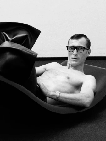 Black and white portrait of a man with glasses, shirtless and reclining on a chaise lounge with a large pillow, indoors.
