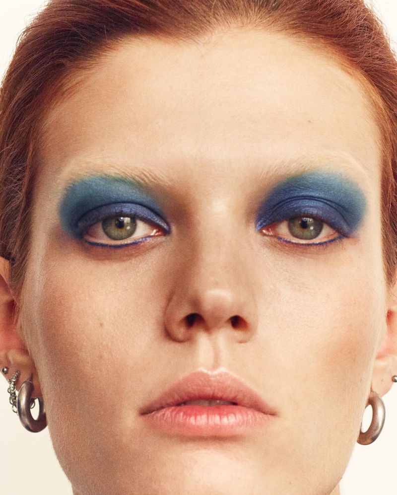 Close-up portrait of a young adult woman with red hair, fair skin, and striking blue eyeshadow on her upper eyelids. She has light green eyes.
