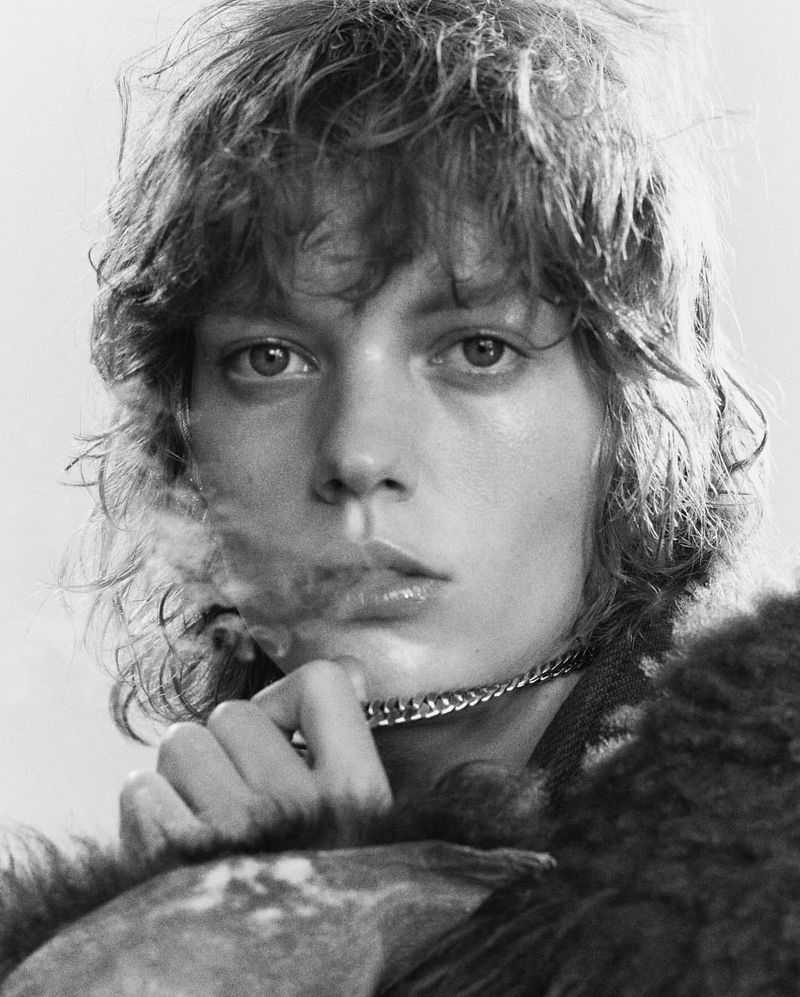 Black-and-white portrait of a young adult woman with curly brown hair, wearing a chain, and holding a fur-like garment in the studio.