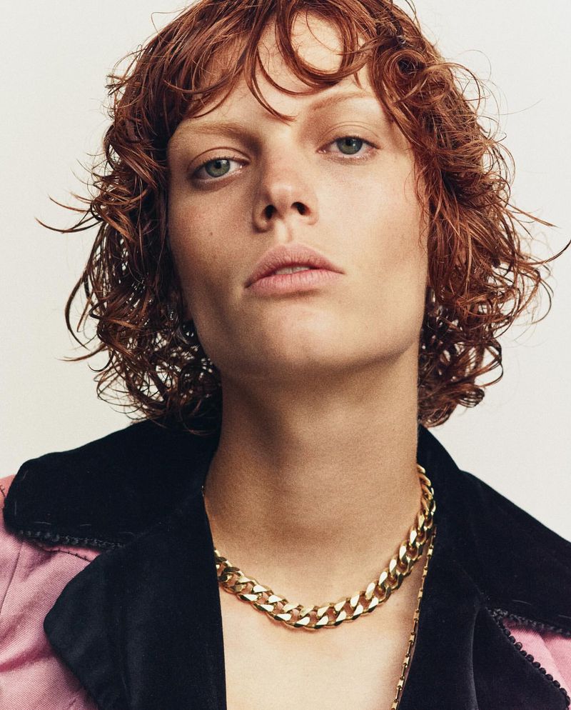 Close-up shot of a fair-skinned adult with auburn curly hair and a gold chain necklace, wearing a pink and black jacket.