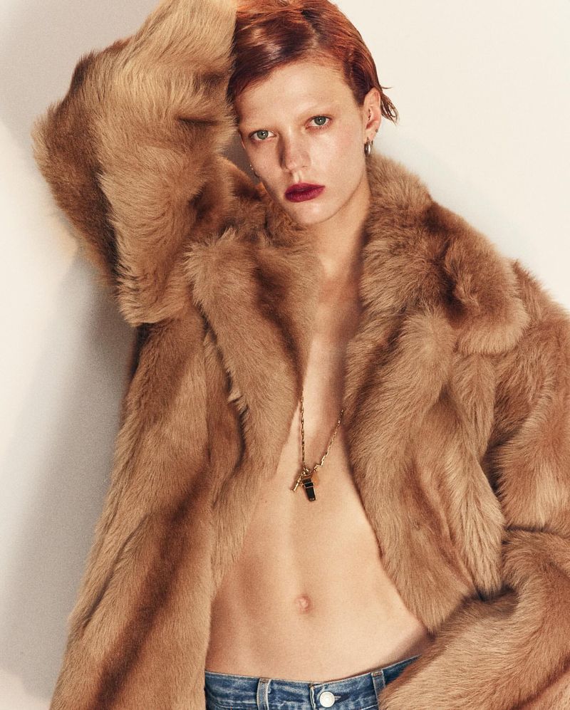 Model with short red hair wearing an open camel fur coat and necklace against a neutral studio backdrop.